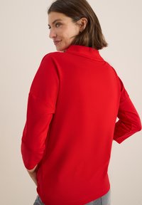 Pull rouge en tissu doux, présentant un col montant et des manches trois-quarts, avec une texture lisse et sans motifs visibles.