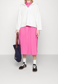 Bílá mikina s kapucí, jasně růžová sukně na knoflíky, bílé ponožky a černé tenisky. Modrá taška přes rameno s grafickým detailem.