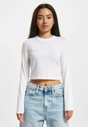 Kuttet langermet topp i hvit bomullstoff, med rund hals og glatt tekstur, kombinert med høytlivs lyseblå jeans.
