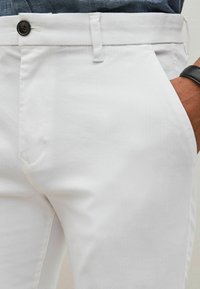 Pantaloni di cotone bianchi con una texture liscia, dotati di un bottone nero, tasche laterali e una vestibilità aderente in vita.