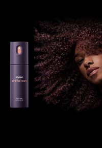 Spray per lo styling multiuso Dyson Chi To San in una bottiglia viola scuro con un pulsante arancione, accanto a capelli testurizzati e ricci.