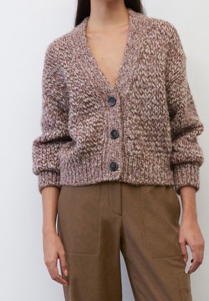 Frau trägt eine grob gestrickte, braune und beige Strickjacke mit Knöpfen, langen Ärmeln und hoch taillierte braune Hose.