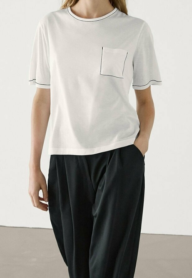 Personne portant une chemise blanche à manches courtes avec bordure noire et détail de poche, associée à un pantalon noir ample, se tenant devant un fond uni.