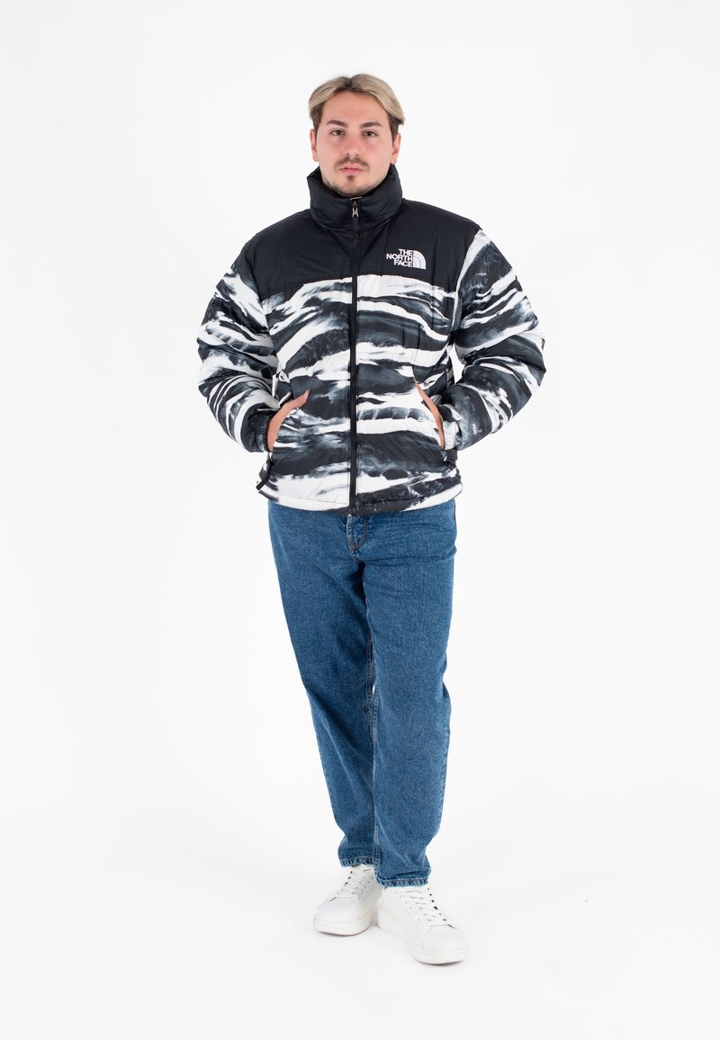 Giacca a maniche lunghe nera e bianca con effetto tie-dye, con colletto alto, chiusura a zip e tasche laterali, abbinata a jeans blu e sneaker bianchi.