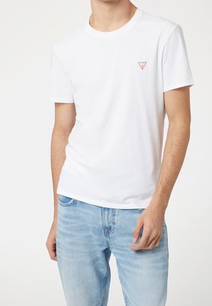 Mężczyzna ubrany w prosty biały T-shirt z krótkim rękawem z małym czerwonym trójkątnym logo na piersi oraz jasnoniebieskie dżinsy, na białym tle.