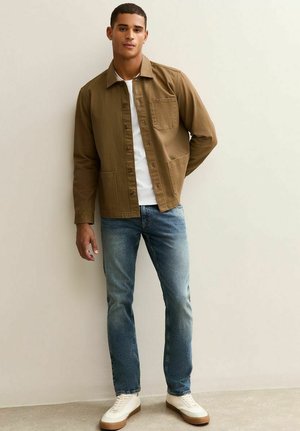 Bruine button-up jasje over een wit shirt, gecombineerd met lichtblauwe jeans en witte sneakers met een bruin zool. Eenvoudig ontwerp, comfortabele pasvorm.