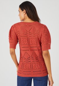Maglione lavorato a maglia in color ruggine con maniche corte e design traforato che presenta motivi geometrici. Tessuto morbido e testurizzato con bordi a costine.