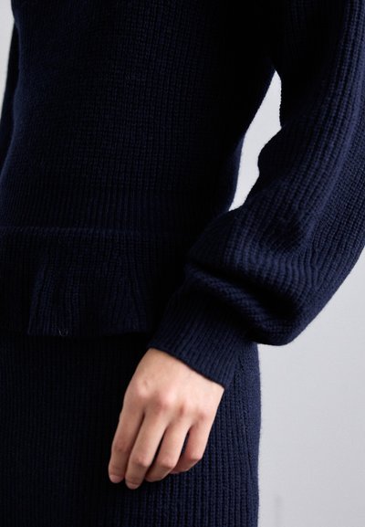 Suéter de lana azul marino con textura de canalé, que presenta un cuerpo ajustado y mangas anchas. Mano descansando suavemente en la cintura, mostrando la profundidad de la tela.