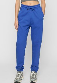 Blå bomullssweatpants med elastisk midje og snøring, med sidelommer og ribbestrikkede mansjetter. Brukt med grå og hvite joggesko.