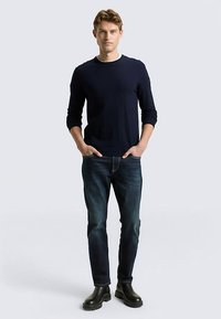 Marineblauer Pullover mit Rundhalsausschnitt, taillierte Silhouette; abgenutzte Jeans mit dunkler Waschung; vervollständigt mit schwarzen Stiefeletten. Glatte Textur insgesamt.
