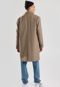 Cappotto lungo in lana marrone con colletto a rib di, design strutturato e polsini con bottoni, abbinato a jeans azzurri chiari e un berretto nero.