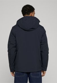 Mørkeblå hættejakke med en glat, mat finish. Har en afslappet pasform, synlige sømdetails og et enkelt design.