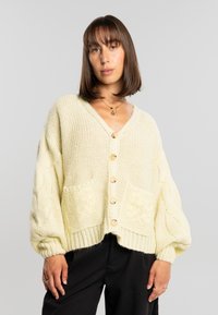 Mujer que lleva un cárdigan de punto grueso color crema con botones de madera y bolsillos delanteros, combinado con pantalones negros y delicados collares dorados.