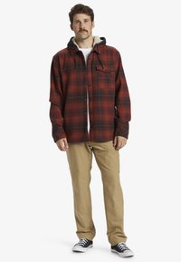 Red and black plaid flannel shirt with a grey hood, featuring a button-up front and a single chest pocket. Worn with khaki pants and sneakers.