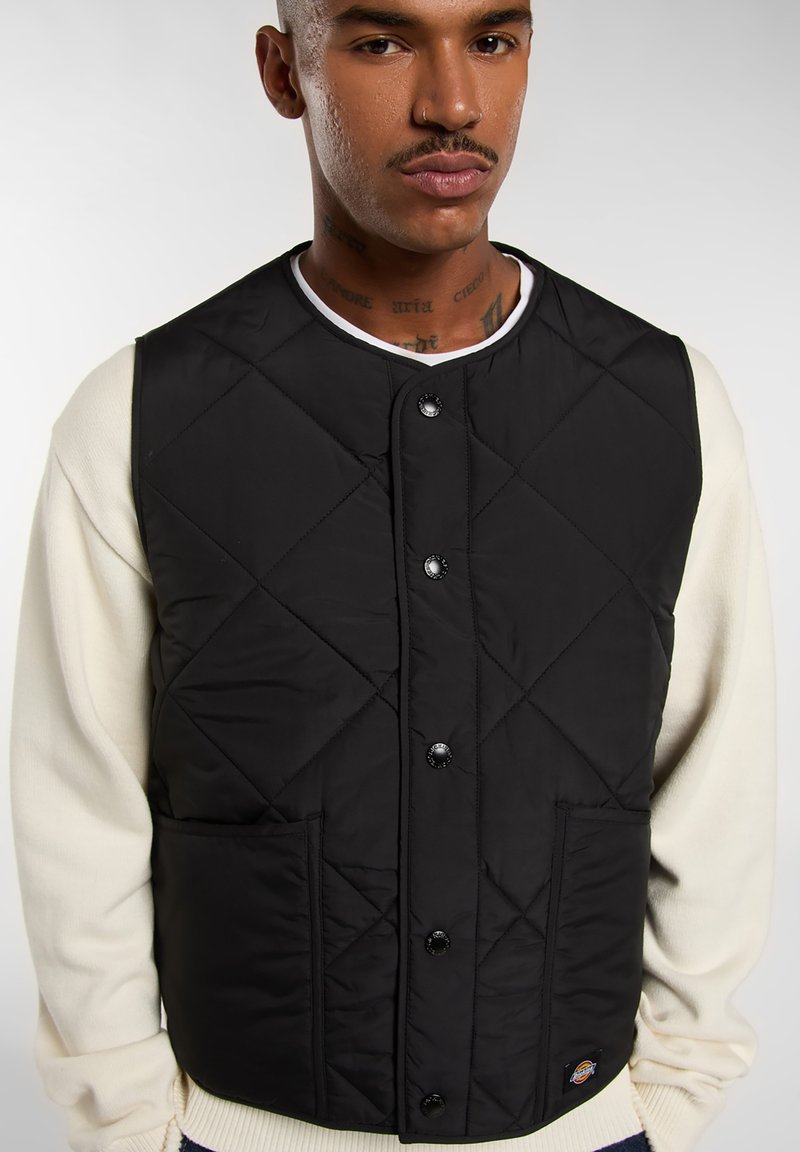 Sort quiltet vest med trykknapper og to forlommer, båret over en hvid crewneck sweater. Har et logo-patch ved kanten.