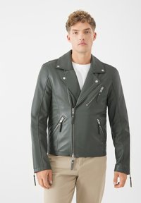 Mörkgrön läderbikerjacka med silverfärgade dragkedjor, flera fickor med dragkedja och en notch-krage. Stylad med en vit skjorta.