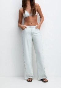 Jean en denim bleu clair à jambes larges avec des boutons décoratifs sur le côté, associé à un haut de bikini triangle assorti. Le modèle pose les mains dans les poches.