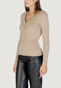 Beige Ripp langärmliges Oberteil mit V-Ausschnitt und drei Knöpfen an der linken Schulter. Kombiniert mit schwarzen Lederhosen. Glatte Textur.