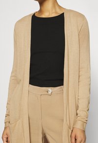 Beżowy kardigan z otwartym przodem, długimi rękawami i bocznymi kieszeniami, noszony na czarnym prążkowanym topie i beżowych spodniach. Miękki, lekki materiał.