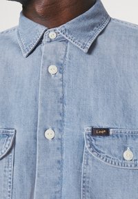 Světle modrá džínová košile s vypasovaným límečkem, dvěma kapsami na prsou a bílými knoflíky. U levé kapsy je malá štítek s nápisem "Lee".