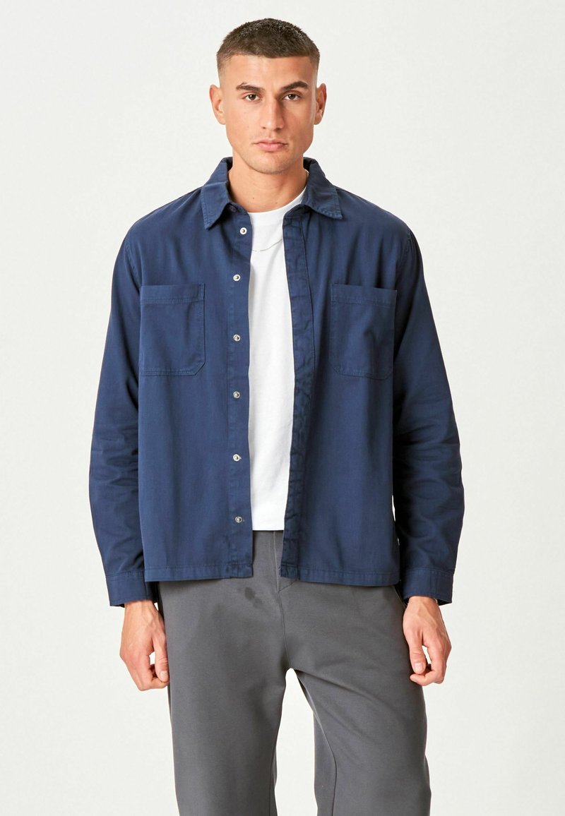 Homme debout portant une chemise à boutons bleu marine par-dessus un t-shirt blanc et un pantalon gris, devant un fond clair uni.
