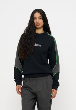 Mlada ženska z dolgimi kodrastimi lasmi, oblečena v črno jopico z zelenimi obrobami in sive hlače, stoji pred enobarvnim ozadjem.