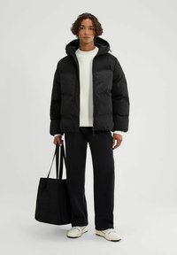 Chaqueta de plumas negra con capucha y cremallera, combinada con un suéter blanco de canalé, pantalones negros y un bolso tote negro. Se puede ver la tela texturizada.