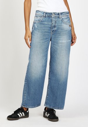 Woman wearing light blue wide-leg jeans and black sneakers with white stripes, standing against a white background.