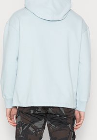 Světlomodrá mikina s volným střihem, vybavená klokáním kapsou a kapucí se šňůrkou, doplněná o maskáčové kalhoty.