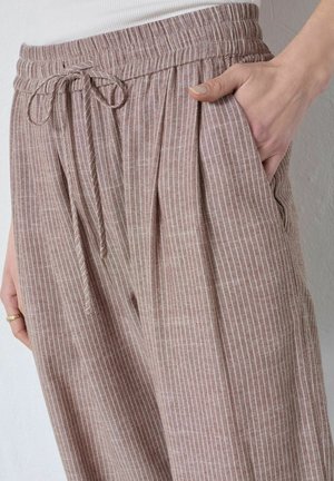 Frau trägt lockere, beige Leinenhose mit weißen vertikalen Streifen und elastischem Bund mit Kordelzug, Hand in der Seitentasche.