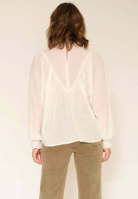 Blouse blanche légère avec des manches longues, design plissé, dos en trou de serrure et couture décorative. Associée à un pantalon vert olive.