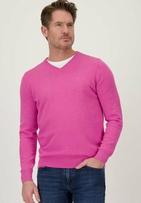 V-hals pullover in felroze gebreid stof met lange mouwen en geribbelde manchetten, gedragen over een wit shirt, gecombineerd met donkerblauwe jeans.