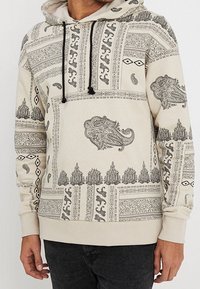 Felpa con cappuccio beige caratterizzata da intricati motivi neri, tra cui disegni paisley e floreali, con cappuccio regolabile e tasca a marsupio frontale.