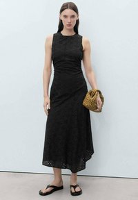 Robe midi noire avec un motif d'œillets floraux, corsage ajusté, jupe évasée et ourlet ondulé. Le mannequin tient un sac à main tissé marron et porte des sandales noires.