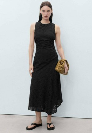 Rochie neagră midi cu un model floral din dantelă, cu bust ajustat, fustă evazată și un tiv ondulat. Modelul ține o geantă din rafie de culoare maronie și poartă sandale negre.