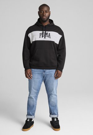 Schwarzer Kapuzenpullover mit weißem Streifen und "PUMA"-Logo, getragen zu hellblauen Jeans und schwarzen Sneakern. Einfaches Design, Material aus Baumwollmischung.