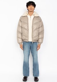Man in een beige pufferjack met een schapenvacht kraag, een crèmekleurige half-zip trui, blauwe flare jeans en donkere schoenen, staande tegen een witte achtergrond.