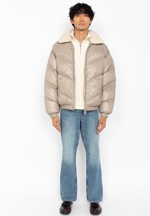 Mand iført beige dunjakke med shearlingkrave, lyscreme halvpullover med lynlås, blå udsvejede jeans og mørke sko, stående foran en hvid baggrund.
