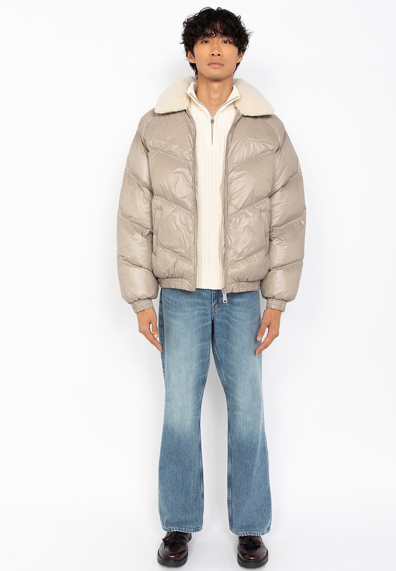 Man in een beige pufferjack met een schapenvacht kraag, een crèmekleurige half-zip trui, blauwe flare jeans en donkere schoenen, staande tegen een witte achtergrond.