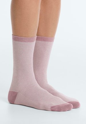 Picioare purtând șosete tricotate texturate, roz deschis, până la jumătatea gambei, cu manșete, călcâie și vârfuri roz mai închis, stând pe o suprafață albă.