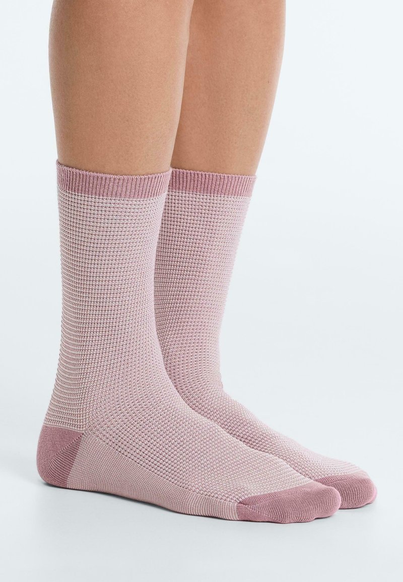 Nohy v ružových pletených ponožkách do polovice lýtok s tmavšie ružovými manžetami, pätami a špičkami, stojace na bielom povrchu.