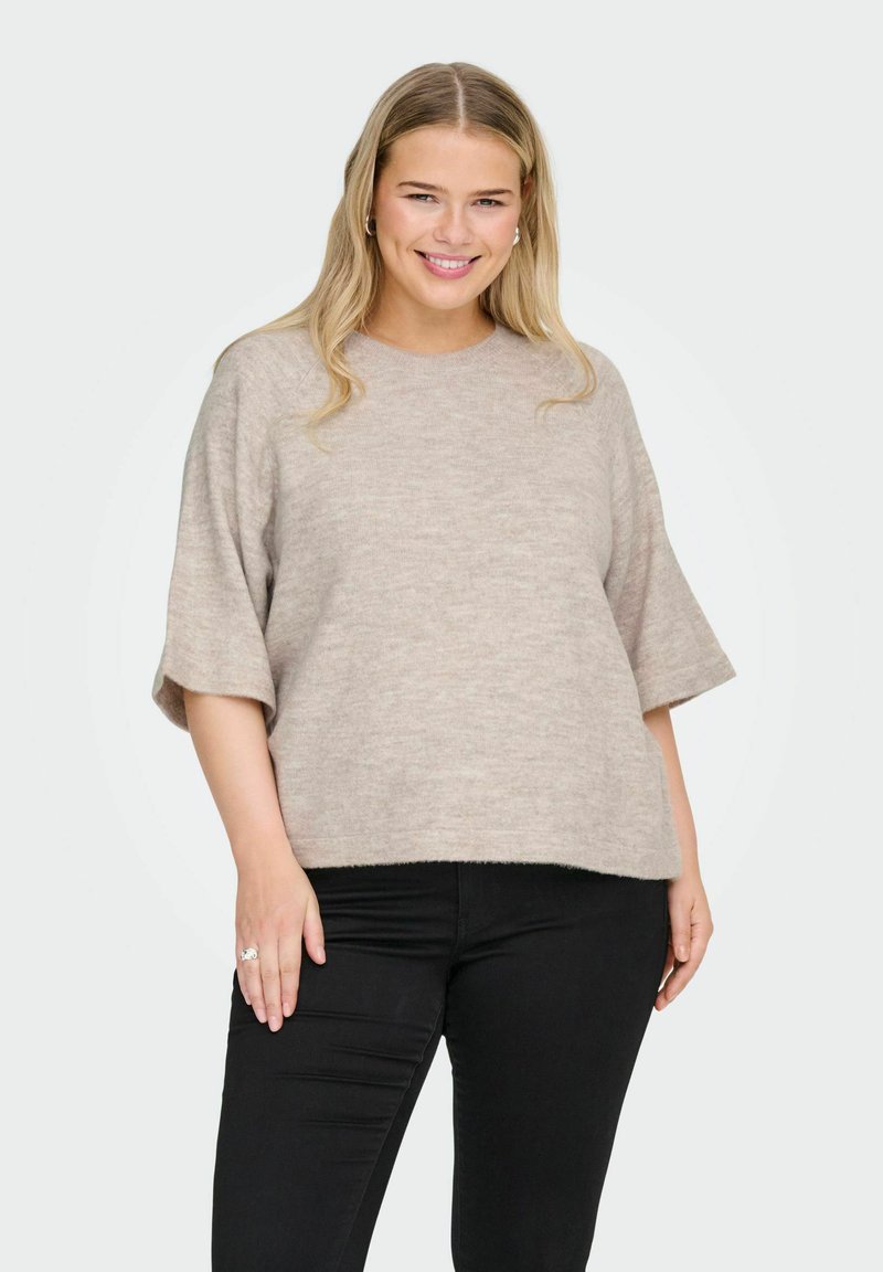 Maglione beige lavorato a maglia con maniche corte e vestibilità rilassata, caratterizzato da una texture morbida e un motivo sottile, abbinato a jeans neri.