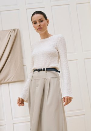 Žena stojaca v interiéri, oblečená v bielom dlhom rukáve s textúrou, béžových nohaviciach s vysokým pásom a širokými nohavicami a čiernym opaskom.