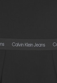 Černá bavlněná látka se širokým pasem, na kterém je horizontálně umístěno "Calvin Klein Jeans" v šedé barvě.