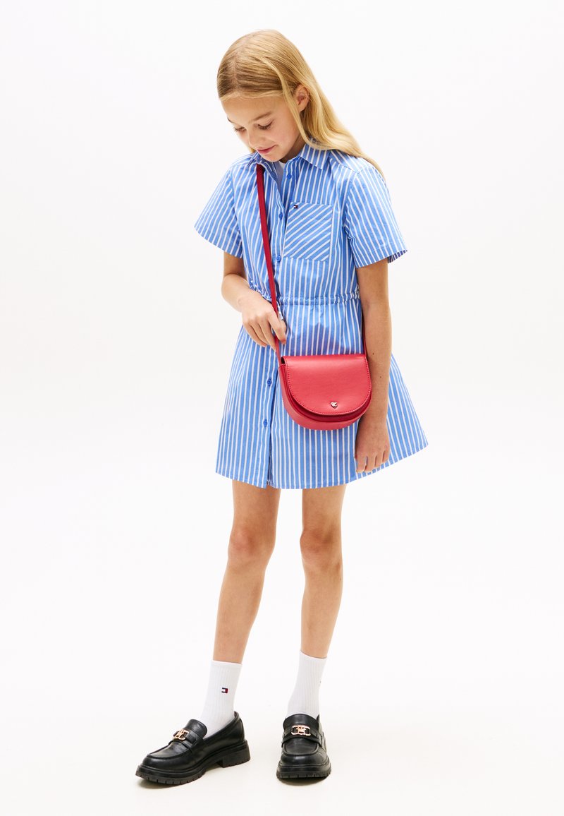 Φόρεμα πουκάμισο με μπλε και λευκές ρίγες, κοντά μανίκια, κούμπωμα με κουμπιά και σφιγμένη μέση. Κόκκινη τσάντα χιαστί και μαύρα παπούτσια με χοντρή σόλα.