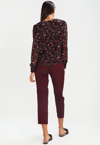 Blusa nera a fiori con maniche lunghe trasparenti, abbinata a pantaloni aderenti bordeaux. La blusa presenta un collo rotondo e un sottile dettaglio con bottoni sul retro.