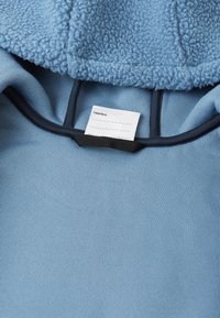 Haut à capuche bleu clair avec une texture douce et duveteuse sur la capuche, et un corps lisse et côtelé. Comprend une étiquette d'entretien à l'intérieur.