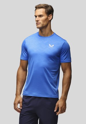Homme portant un t-shirt de sport bleu ajusté à manches courtes avec un logo ailé blanc sur la poitrine, regardant sur le côté sur un fond uni.
