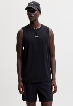 Homme portant un débardeur de sport noir, un short noir et une casquette noire, debout devant un fond clair uni.