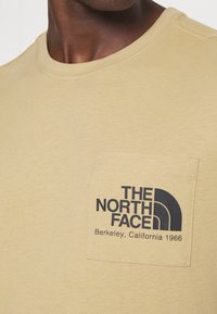 Zblízka na béžovou kapsu trička s černým logem "The North Face Berkeley, California 1966" na hrudi.
